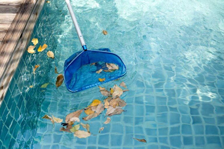 A pool cleaning pole scoops up a pile of leaves sitting on top of a beautiful blue pool.