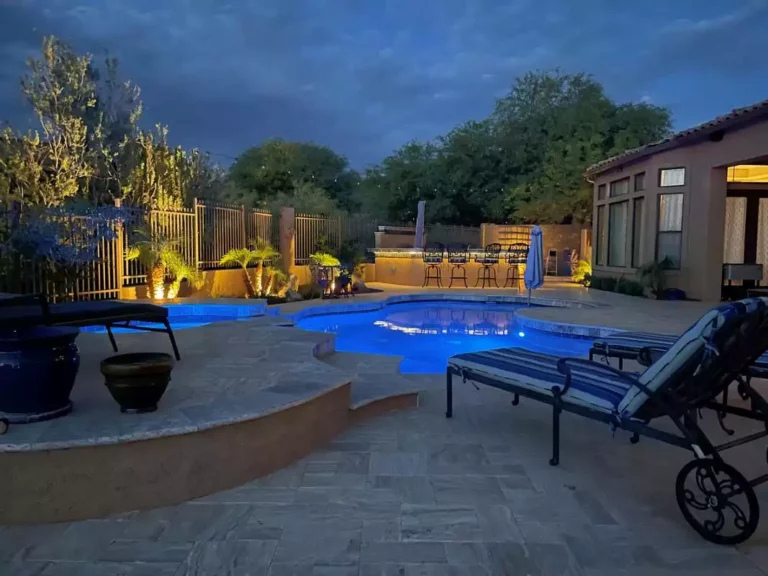 A backyard pool with blue lighting in Arizona in the evening, surrounded by a patio and desert trees.