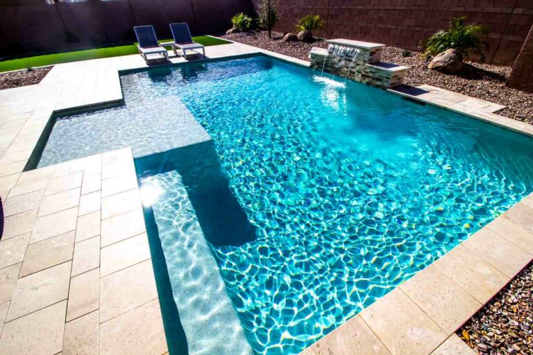 A view of a backyard sunny swimming pool featuring a waterfall and two lounge chairs.