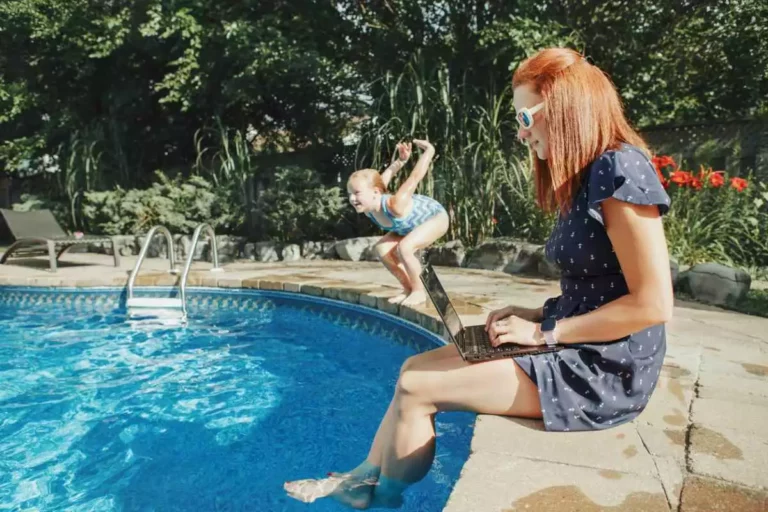 A child winds up to jump into the pool as her mom sits on the edge working on her laptop.