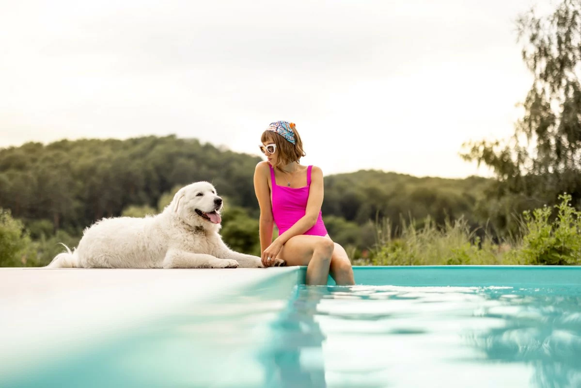 A red-headed young woman in a pink swimsuit sits poolside with her fluffy, white dog.